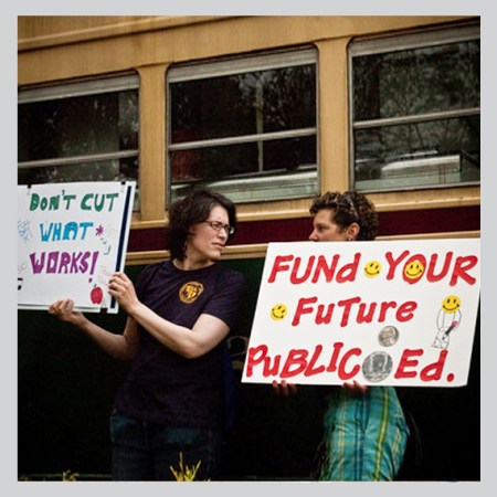 People stand in front of a school bus and protest public school funding cats with posters.