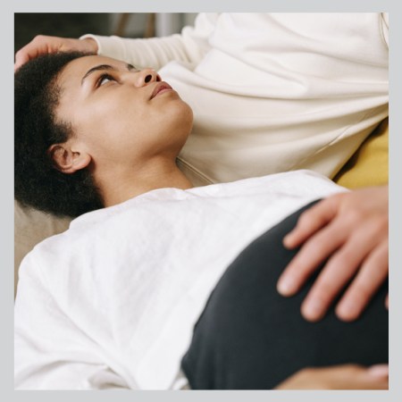 black woman lies on her back looking pensive with another's hand on her belly